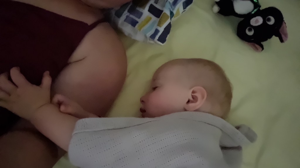 Late-night photo of baby sleeping next to mother, with little toddler hand on mum's breast, both covered in a cuddle blanket in bed.